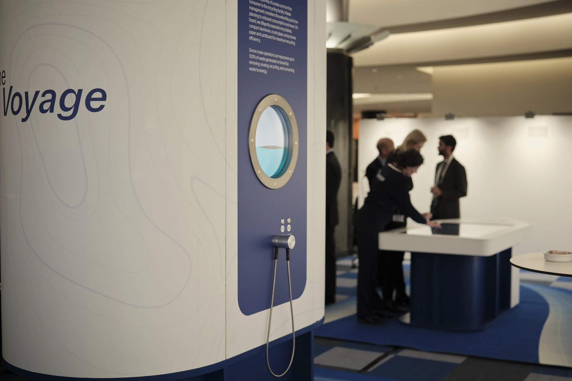The Voyage installation, a curved white shell with a porthole screen set into it, with guests at the touchscreen table behind