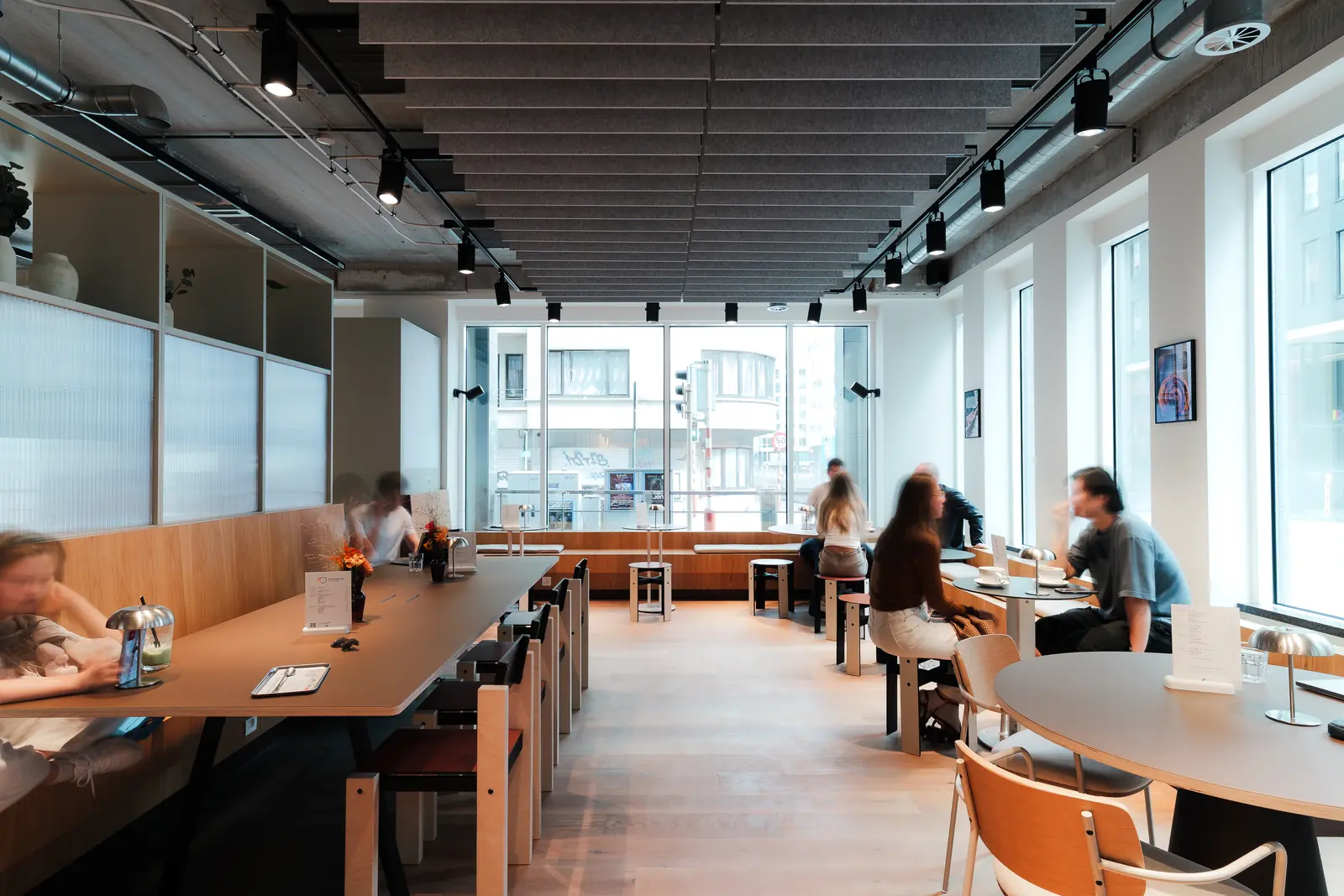 Guests working and talking along the long communal tables, with daylight from the street windows