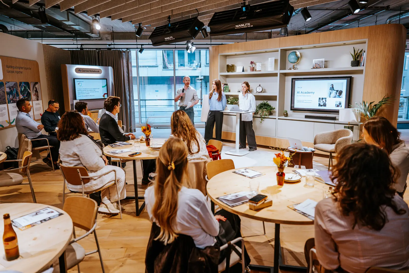 A talk in progress, speakers standing while the audience listens from round tables